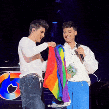 a man holding a rainbow flag next to another man holding a microphone with urrumagics written on the bottom right