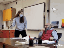 a man sits at a desk in front of a white board while a woman stands behind him wearing a mask