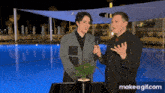 a man is holding a microphone and talking to another man in front of a swimming pool