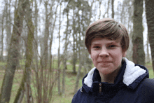 a young man in a blue jacket with a white collar stands in the woods