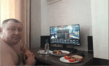 a shirtless man is sitting at a table with a computer monitor and a plate of food .