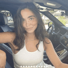 a woman in a white tank top sits in the driver 's seat of a car