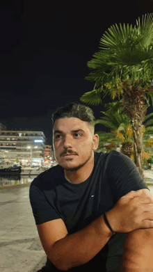 a man in a black t-shirt sits in front of a palm tree at night