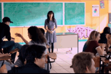 a woman stands in front of a group of people in a classroom with graffiti on the wall behind her