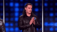 a man in a leather jacket is clapping his hands while standing in front of a blue background .