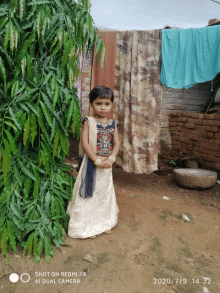 a little girl is standing in front of a brick wall and a tree with a shot on redmi y3 ai dual camera