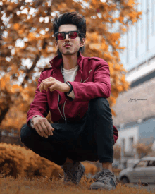 a man wearing sunglasses and a red jacket is squatting down in the grass