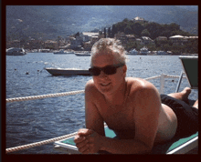 a shirtless man wearing sunglasses is laying on a beach chair in front of a body of water