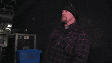 a man wearing a black hat and a purple plaid shirt stands in a dark room