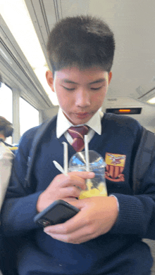 a boy in a school uniform looks at his phone while holding a drink