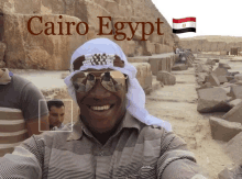 a man wearing sunglasses and a head scarf is smiling in front of a cairo egypt sign