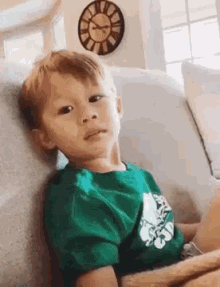 a young boy in a green shirt is sitting on a couch with a clock on the wall behind him .