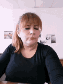 a woman with red hair and a black shirt is sitting at a desk in front of a wall with pictures on it