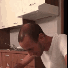 a man in a white shirt is sitting at a table in a kitchen with a sink and a stove .