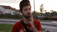 a man with a beard wearing a red shirt and a black hat is standing in front of a street .