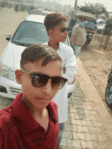 a boy wearing sunglasses stands next to another boy in front of a white car