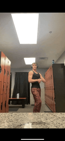 a man in a black tank top and plaid pants stands in a gym locker room