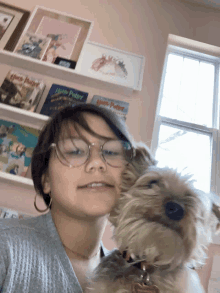 a girl with glasses holds a small dog in front of a bookshelf with harry potter books