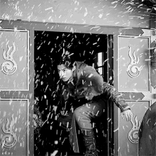 a black and white photo of a man standing in a doorway with snow falling and the letters k & d on the bottom right