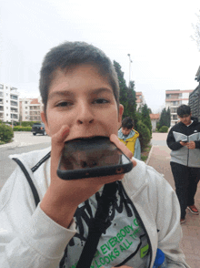 a boy wearing a shirt that says " everybody looks all the time " holds a cell phone in his mouth