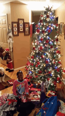 a girl in a pony shirt stands in front of a decorated christmas tree