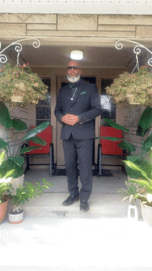 a man in a suit stands in front of a porch with potted plants