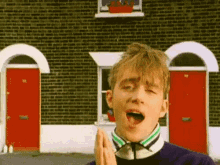 a young man is standing in front of a brick building with his mouth open and his hands folded in front of him .