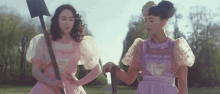 two girls in pink dresses are standing next to each other holding shovels in a field .