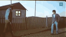 two men are standing next to each other in front of a wooden fence in front of a house .