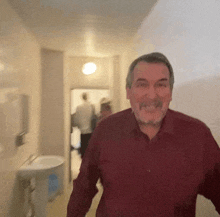 a man in a red shirt is walking down a hallway with a sink in the background .