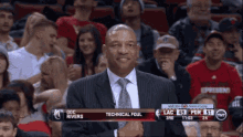 a man in a suit and tie stands in front of a crowd at a basketball game with the name doc rivers on the screen