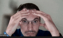 a man wearing glasses holds his hands to his forehead in front of a screen that says iskewycz