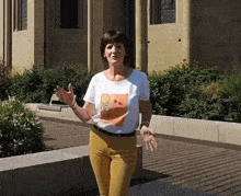 a woman in a white t-shirt and yellow pants is standing in front of a building with her arms outstretched .