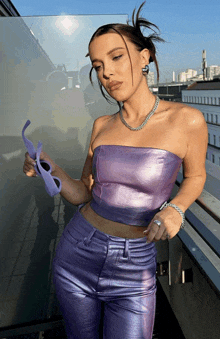 a woman wearing a purple top and purple pants holds a pair of sunglasses