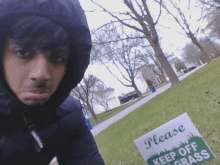 a man wearing a hooded jacket is standing next to a sign that says please keep off grass