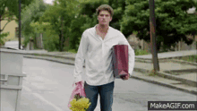 a man in a white shirt is walking down the street holding a bag of flowers and a gift bag .