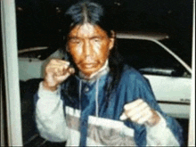 a man with long hair and a blue jacket is standing in front of a car .