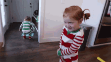 a little girl in a red and white striped pajama stands next to a toddler in a green and white striped pajama