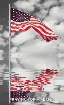 a black and white photo of an american flag waving in the wind and its reflection in the water .