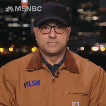 a man wearing glasses and a jacket with velshi embroidered on it