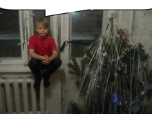 a young boy in a red shirt sits on a window sill next to a christmas tree