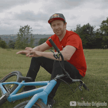 a man in a red shirt is sitting next to a blue bicycle with youtube originals written on the bottom