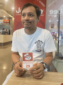 a man in a jollibee t-shirt sits at a table holding a sign that says 29