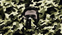 a man with a mask on his face is behind a camouflage cloth