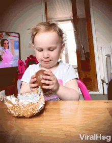 a little girl sitting at a table holding a chocolate egg with the word viralhog on the bottom right corner