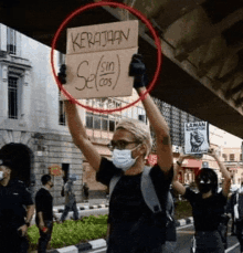 a woman wearing a mask is holding a sign that says ' kerajaan se sin cos ' on it .