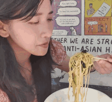 a woman eating noodles with chopsticks in front of a sign that says " her we are strong anti-asian "