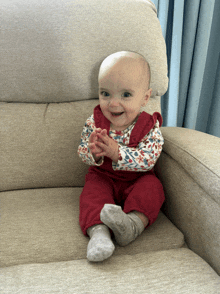 a baby in red overalls sits on a couch and claps her hands