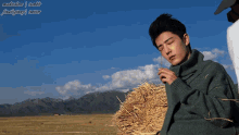 a man in a green sweater is standing next to a pile of hay with mountains in the background ..