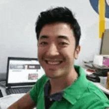 a man in a green shirt is smiling while sitting at a desk with a laptop .